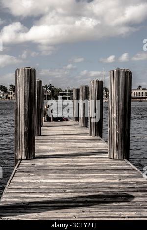 Persona seduta sul molo di legno che si estende in acque calme vicino alle case costiere Foto Stock
