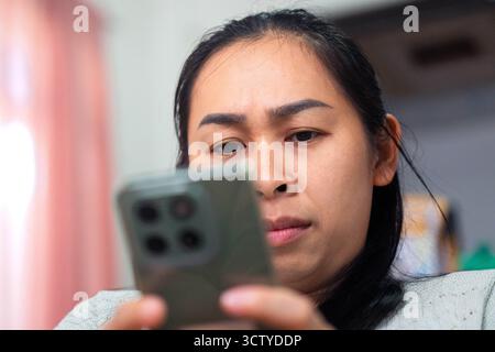 Primo piano di una donna asiatica che soffre di affaticamento agli occhi mentre guarda uno smartphone. L'immagine rappresenta l'affaticamento digitale, l'uso eccessivo dello schermo e la visione p Foto Stock