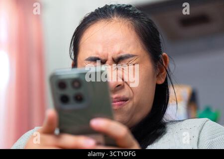 Primo piano di una donna asiatica che soffre di affaticamento agli occhi mentre guarda uno smartphone. L'immagine rappresenta l'affaticamento digitale, l'uso eccessivo dello schermo e la visione p Foto Stock