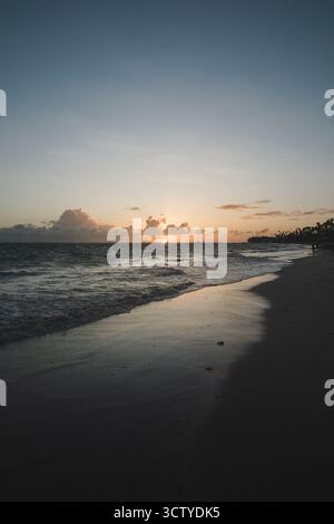 Onde dolci si infrangono sulla riva mentre il sole tramonta all'orizzonte, generando colori caldi sul cielo. La tranquilla spiaggia invita a riflettere tranquillamente al crepuscolo Foto Stock