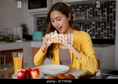 Donna asiatica adulta gode di una sana colazione a casa. Mangiare un panino, sorseggiare succo di frutta e mele in una cucina moderna. Una mattinata allegra. Foto Stock