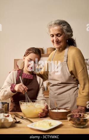 Donna anziana caucasica accanto alla ragazza caucasica che sbatte la pastella in una ciotola di vetro, entrambi indossano grembiuli, preparano il cibo insieme in cucina, sorridono e interagiscono Foto Stock
