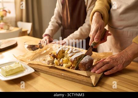 Donna caucasica di mezza età che affetta la torta appena sfornata su una tavola di legno, mentre una donna caucasica anziana in piedi nelle vicinanze assiste in cucina, mani visibili, burro sul piatto in primo piano Foto Stock