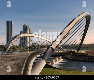 Ponte pedonale sul lago a Taman seri Empangan, Putrajaya, Malesia. Moderno design ad arco su cascate, con grattacieli contemporanei visibili sotto un cielo blu limpido. Foto Stock