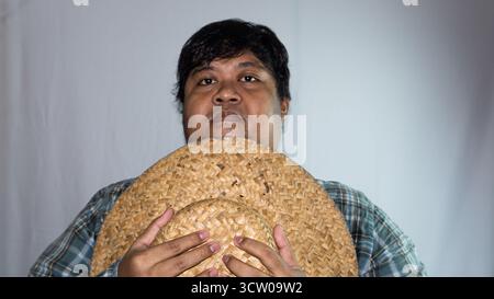 Uomo indonesiano che indossa un cappello di paglia e una camicia a scacchi verde su sfondo bianco. Cappello di paglia tenuto in mano Foto Stock