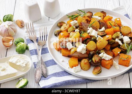 calda insalata autunnale di zucca arrosto, patate, cavoletti di bruxelles cosparsi di feta sbriciolata su un piatto su un tavolo di legno bianco, vista orizzontale fr Foto Stock