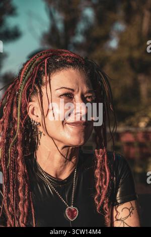 ritratto di una donna matura con dreadlock rossi e verdi che indossano un top da motociclista nero lucido e una collana a forma di cuore rosso Foto Stock
