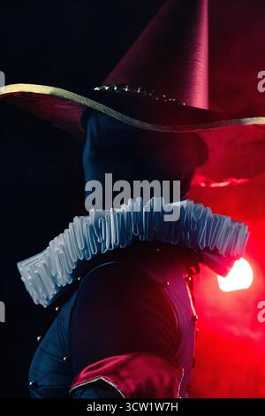 Spettacolare ritratto ravvicinato e dall'angolo basso di un misterioso giullare o di un personaggio di harlequin con cappello conico e colletto bianco con ruff su sfondo scuro Foto Stock