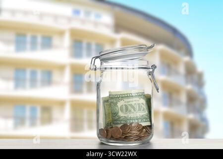 Barattolo con dollari e monete contro il condominio. Risparmiare denaro per comprare un appartamento Foto Stock