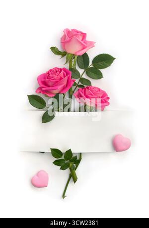 Fiori rosa rose, cuori e carta bianca con spazio per testo su sfondo bianco. Vista dall'alto, disposizione piatta Foto Stock