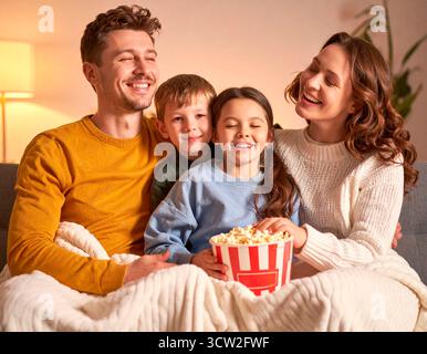 Allegra famiglia che rideva e mangiava popcorn mentre sedeva sul divano sotto una coperta Foto Stock