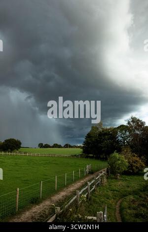 Una vista spettacolare delle nuvole di tempesta scure che si radunano sopra campi verdi e recinzioni di legno nella campagna inglese. L'immagine cattura la calma solo prima Foto Stock