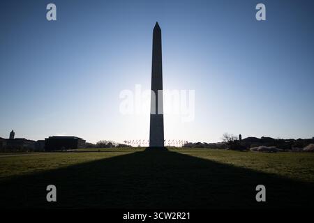 Washington Monument dal National Mall Washington DC | WASHINGTON D.C., Stati Uniti: Il Washington Monument si erge in sagoma contro un cielo limpido, con la sua ombra che si estende attraverso il National Mall verso lo Smithsonian's National Museum of African American History and Culture. Progettato da Robert Mills e completato nel 1884 dopo 23 anni di costruzione, l'obelisco in marmo si innalza di 555 piedi 5⅛ pollici (169,29 m) e rimane la struttura in muratura più alta del mondo. Un anello di cinquanta bandiere americane, una per ogni stato, circonda la sua base. Foto Stock