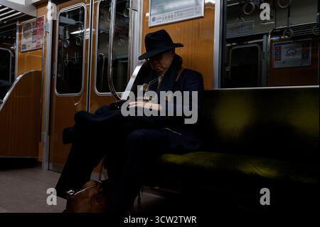 Un gentiluomo con un cappello e una tuta scura riposa tranquillamente su un treno della metropolitana di Osaka, illuminato da una luce soffusa dell'interno Foto Stock