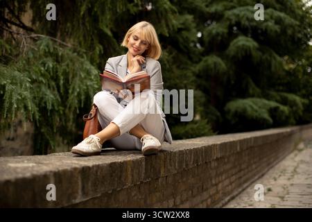 Una donna siede su un muro di pietra, impegnata a leggere un libro. Appare rilassata e premurosa, circondata da alberi verdi in un ambiente all'aperto sereno Foto Stock