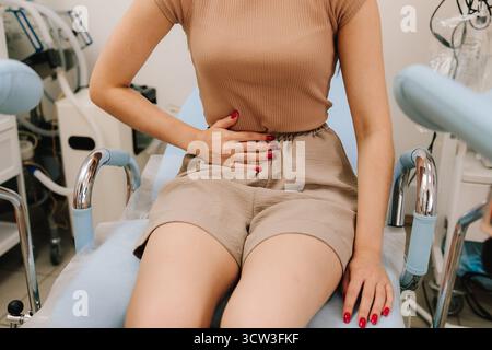Una giovane donna siede su una sedia ginecologica tenendo l'addome in dolore. Il fastidio addominale durante la consultazione segnala possibili problemi ginecologici. W Foto Stock