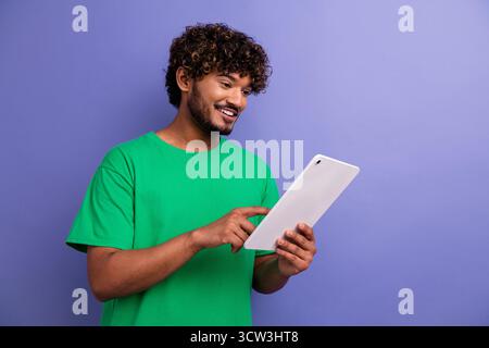 Il giovane con i capelli ricci usa un tablet sorridente su uno sfondo viola e indossa una t-shirt verde, sottolineando la tecnologia e il tempo libero Foto Stock