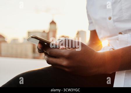 Vista ravvicinata dell'irriconoscibile uomo nero che invia messaggi sullo smartphone mentre è seduto al tramonto all'aperto Foto Stock