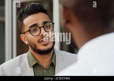 Vista a spalla di un giovane e bell'uomo barbuto mediorientale con occhiali che parla con un collega maschile irriconoscibile all'aperto Foto Stock