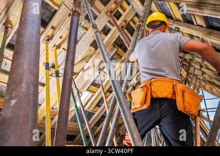 Un operaio edile che indossa un casco e un'imbracatura monta un ponteggio in un sito alto, concentrandosi sulla sicurezza e la precisione durante il giorno. Foto Stock