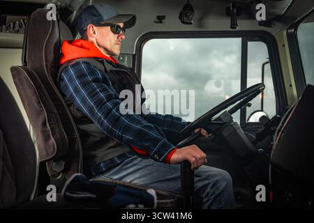 Un conducente esperto siede in sicurezza nella cabina di un camion pesante, indossando occhiali da sole e un berretto, mentre naviga attraverso un ambiente nuvoloso. Foto Stock