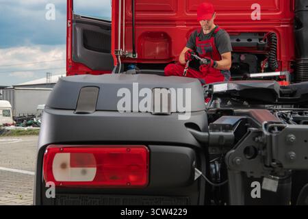 Il meccanico in tuta rossa lavora su un camion in un garage, utilizzando gli attrezzi per riparare un pezzo mentre è circondato da veicoli, concentrandosi sul suo compito. Foto Stock