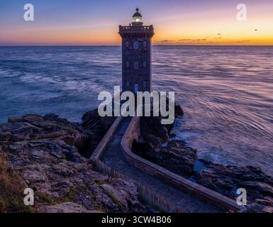 Un faro solitario, il faro di Kermorvan, si erge a guardia sulla costa frastagliata, bagnato dalle calde sfumature del sole che tramonta. Il percorso verso il lightho Foto Stock