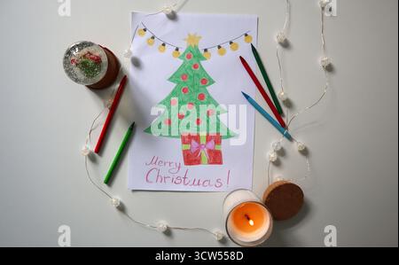 Albero di Natale disegnato a mano con ornamenti rossi e confezione regalo, circondato da matite colorate, globo di neve, candela e luci delle fate. Accogliente appartamento per le feste Foto Stock