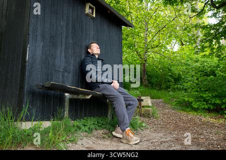 Uomo in un momento tranquillo sulla panchina nella foresta goditi la natura, uomo appoggiato contro un capannone di legno felice in un momento naturale Foto Stock