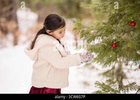 Una ragazza con comodi abiti invernali ammira gli ornamenti rossi su un pino innevato Foto Stock