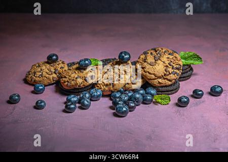 Un delizioso trio: Biscotti al cioccolato appena sfornati, un frullato freddo di mirtilli e banane e una ciotola di mirtilli freschi su un morbido e minimalista pu Foto Stock