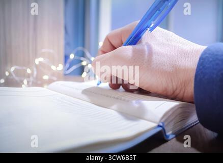 Un uomo scrive i suoi piani e obiettivi per il nuovo anno in un quaderno, mentre le piccole ghirlande brillano nelle vicinanze Foto Stock