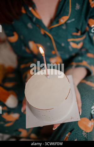 Festeggiamo il compleanno a casa, una ragazza che tiene la torta con la candela. Una ragazza in pigiama tiene una torta bianca fatta in casa con una candela in fiamme Foto Stock