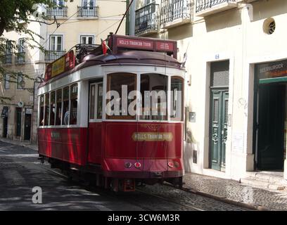 Tram rosso su strada acciottolata a Lisbona, Portogallo - Un tram turistico rosso d'epoca (Hills Tramcar Tour) che si sposta lungo una strada acciottolata incorniciata dalla luce Foto Stock