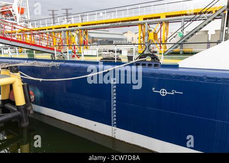 Un vivace ambiente portuale mostra una grande nave cisterna blu ormeggiata al molo, con tubature gialle e piattaforme industriali che creano una logistica dinamica Foto Stock