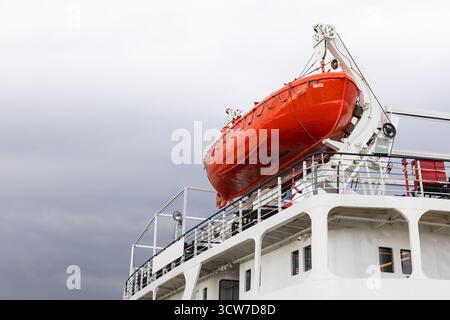 Scialuppa di salvataggio arancione fissata sul ponte di una grande nave bianca, adagiata contro un cielo nuvoloso grigio. Questa immagine trasmette sicurezza, prontezza al salvataggio e navigazione Foto Stock