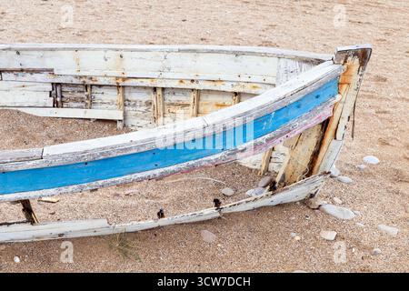 Un'antica barca battuta dal tempo si trova su una spiaggia sabbiosa, con la sua striscia blu che si stacca. Chiodi arrugginiti, legno grezzo e pietre sparse evocano nostalgia, solit Foto Stock