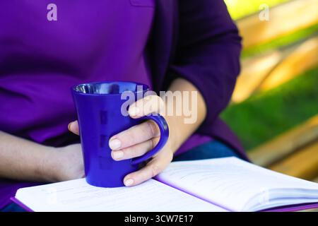 donna che legge il tazzino del libro tè viola fuori dalla natura del parco. una donna o una donna che legge un libro all'aperto in un parco o in un giardino, tenendo in mano una tazza di tè, con un hin Foto Stock