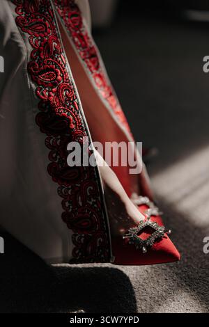 Orlo dell'abito da sposa Vyshyvanka ricamato rosso e moderna scarpa rossa con tacco alto e fibbia in cristallo, impreziosita dalla luce del sole e dall'ombra. Autentica etnia f Foto Stock