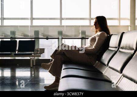 Una donna siede su una panchina nera in una sala d'attesa. Indossa una camicia bianca e pantaloni abbronzati Foto Stock