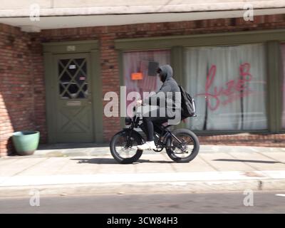 Ciclista elettrica che si muove lungo un marciapiede indossando una felpa con cappuccio nera e sneakers bianche. Il passaggio di piccole imprese costituisce un pericolo per gli escursionisti. Foto Stock