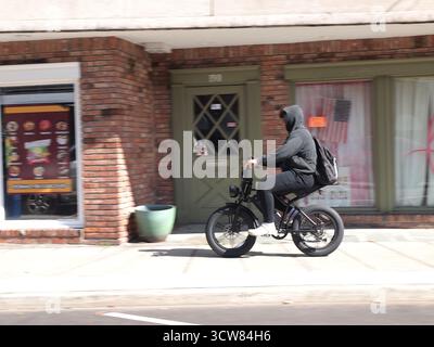 Ciclista elettrica che si muove lungo un marciapiede indossando una felpa con cappuccio nera e sneakers bianche. Il passaggio di piccole imprese costituisce un pericolo per gli escursionisti. Foto Stock