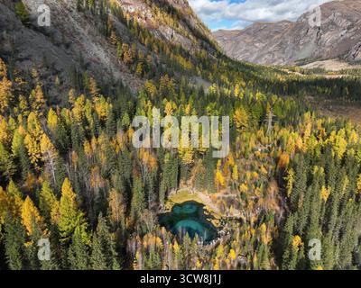Una splendida vista aerea di un piccolo e vivido lago geyser turchese circondato da una vibrante foresta autunnale e da montagne rocciose Foto Stock