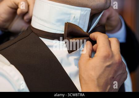 Uno scatto ravvicinato raffigura le mani che regolano un papillon marrone su uno sposo indossando una camicia bianca e un gilet scuro durante i preparativi per il matrimonio, catturando l'eleganza Foto Stock
