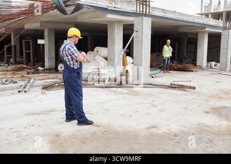 Costruttore che indossa un elmetto in piedi sul cantiere per controllare l'orologio da polso mentre il giovane uomo Nero in un giubbotto di sicurezza in piedi sullo sfondo osservando i progressi Foto Stock