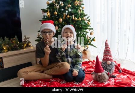 Due ragazzi allegri nei cappelli di Babbo Natale che mangiano biscotti al cioccolato di pan di zenzero vicino all'albero di Natale decorato con ornamenti dorati. Concetto di chicche per le vacanze, f Foto Stock