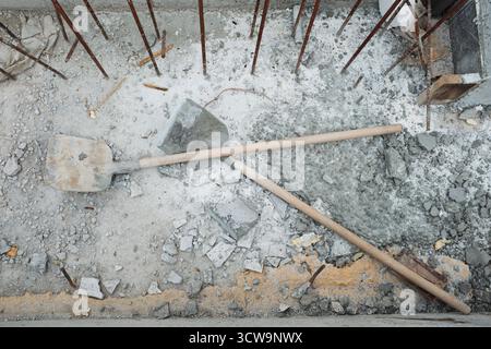 Cantiere con pala e manico in legno adagiati su un pavimento in cemento con detriti sparsi e tondini in metallo esposti, illustrando il processo di costruzione e gli attrezzi di lavoro manuali Foto Stock