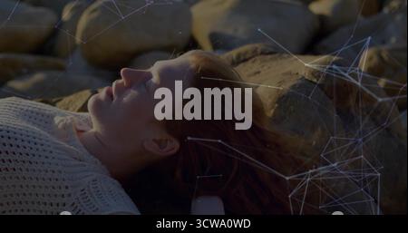 Donna sdraiata che indossa la parte superiore bianca sulla costa rocciosa, con rete luminosa e contenitore sotto i capelli Foto Stock