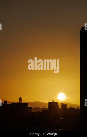 La vivace alba dipinge il cielo mattutino con tonalità dorate, mescolando delicatamente i colori mentre il brillante globo del sole si libra sulle deboli e lontane colline dietro la città dalla silhouette sagomata. Foto Stock