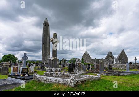 La Torre Rotonda di Kilmacduagh e le croci celtiche in un cimitero con le rovine della cattedrale al monastero Kilmacduagh, Gort, Galway, Irlanda, sotto nuvole grigie. Foto Stock